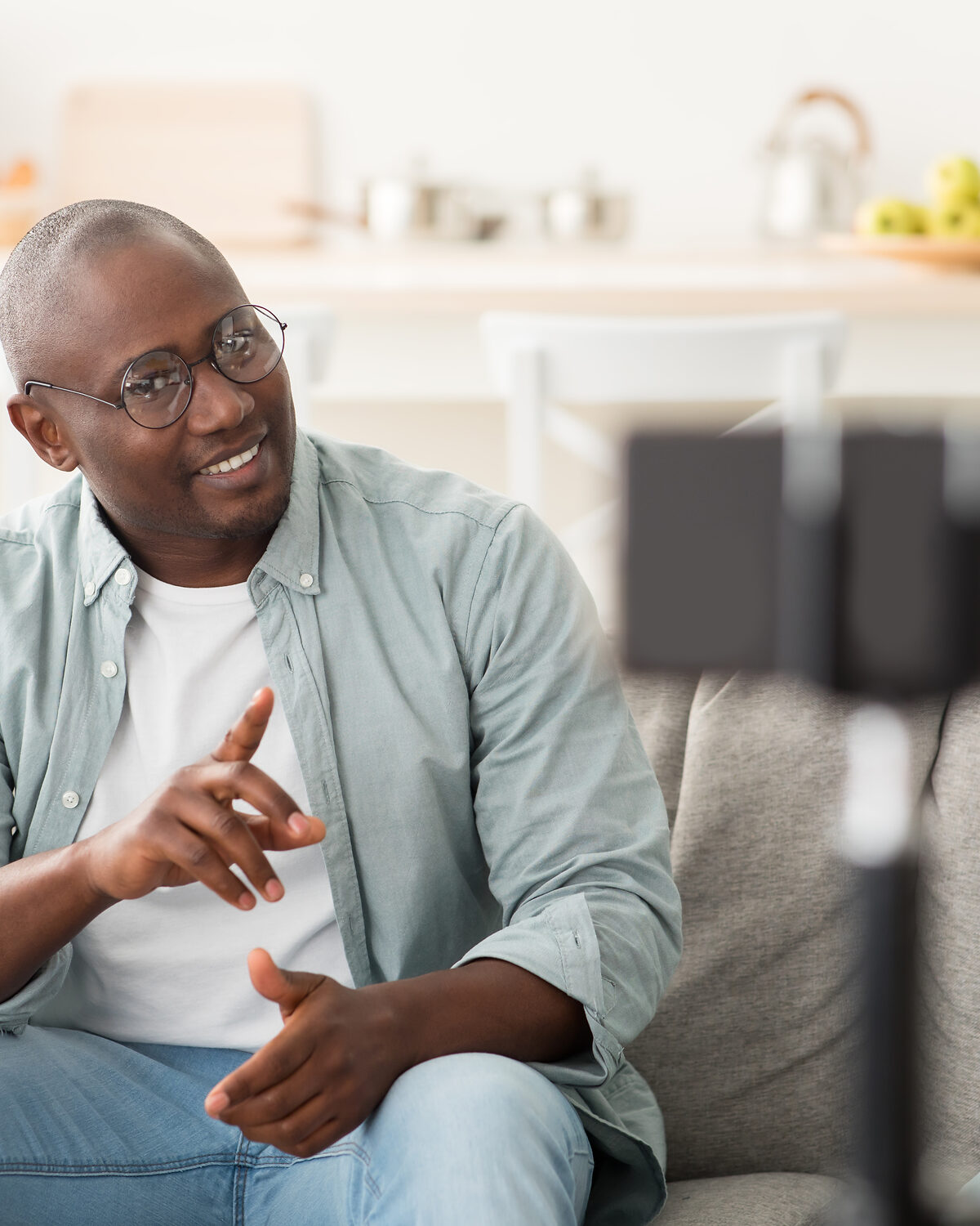 A man sitting on a couch recording himself with a camera, in a thoughtful posture.